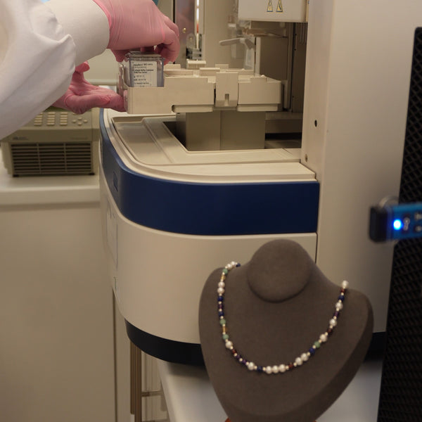 A gloved hand loading a DNA sample into the genetic analyser, with a finished DesigNA necklace beside it